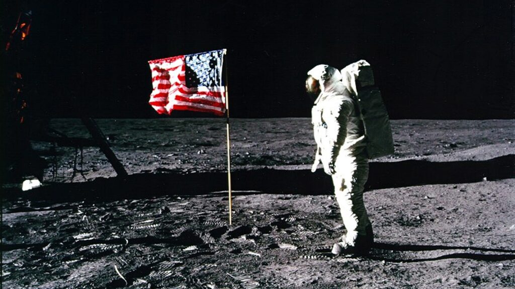 Color photograph taken on the surface of the Moon during the Apollo 11 mission in July 1969. It shows an astronaut in a white extravehicular mobility unit standing near a planted United States flag on the lunar surface. The ground is gray and dusty, marked by footprints and equipment tracks, with the black sky of space in the background. The image was captured at the Sea of Tranquility (Mare Tranquillitatis) shortly after the first human landing on the Moon.