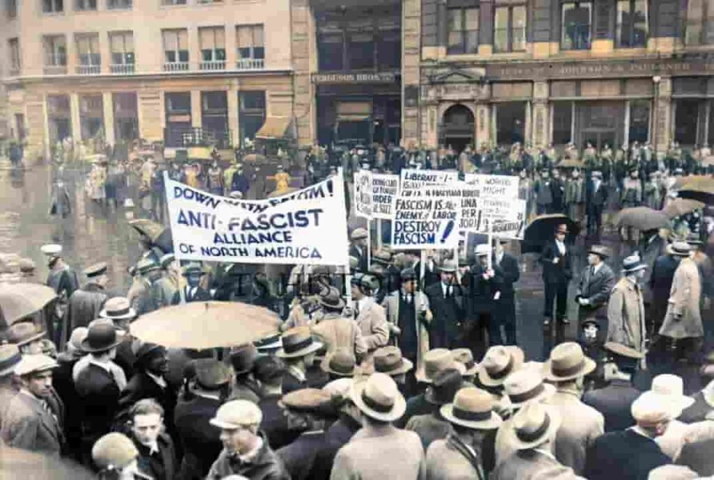 Black and white historical photograph of a large crowd gathered in a city street during the early to mid twentieth century. People wear period clothing including hats and coats and hold protest banners reading “Anti Fascist Alliance of North America” and slogans opposing fascism. The crowd fills the foreground, with buildings and additional demonstrators visible in the background.