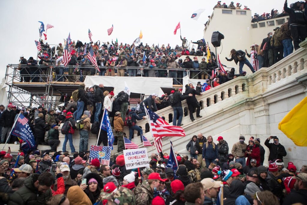 Color photograph taken on January 6, 2021, showing a large crowd gathered on the steps and terraces of the United States Capitol. Many people wear winter clothing and hats and hold American flags and political banners. Some individuals are climbing railings and scaffolding, while others stand densely packed below. The scene conveys a chaotic, crowded protest atmosphere around the Capitol building.